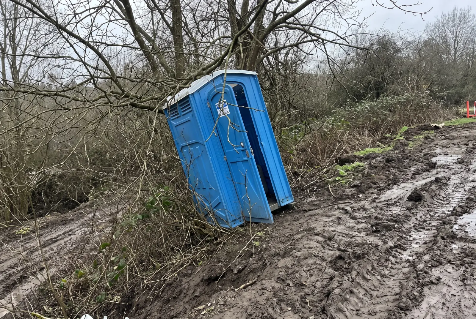 Portable toilet poorly placed on a muddy slope blocked by low tree branches